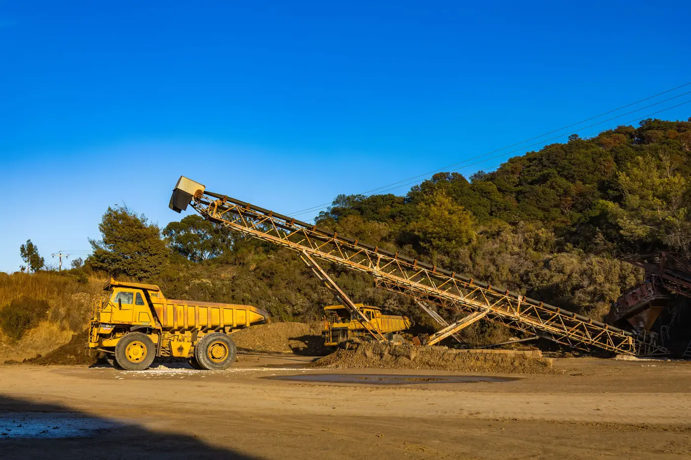 Yellow haul trucks beside an inclined overland conveyor stacker piling material at a mine site.