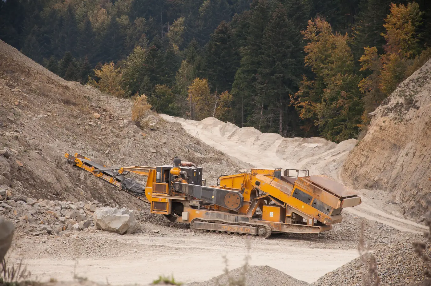 A yellow rock crusher machine on a gravel quarry track between piles of crushed stone.
