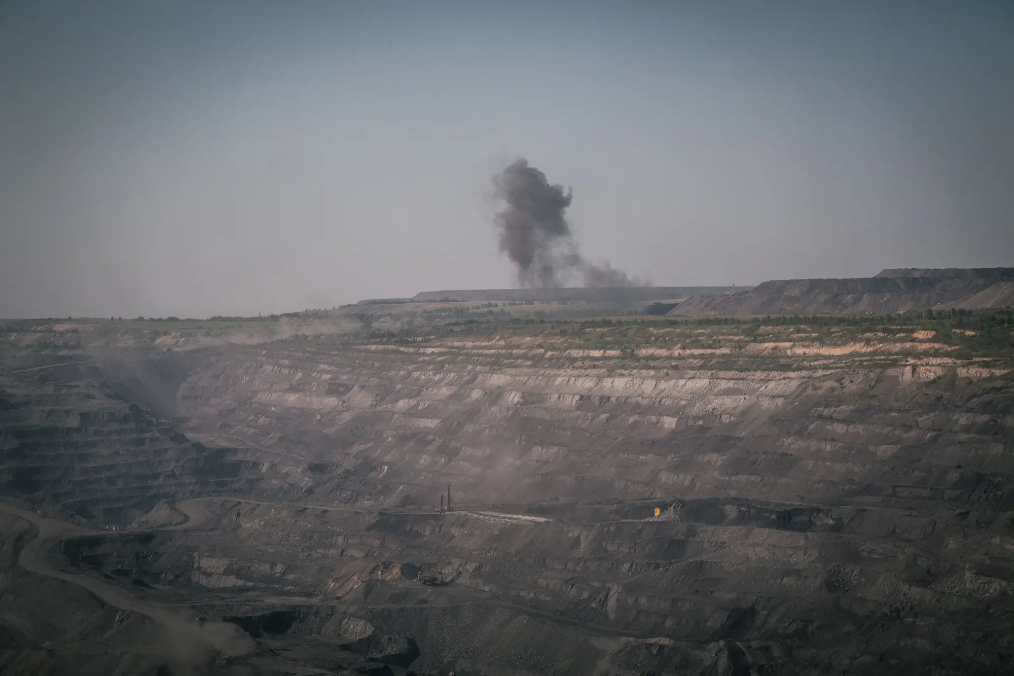 Terraced benches of an open-pit coal mine seen from a distance, with a plume of blast smoke on the far rim